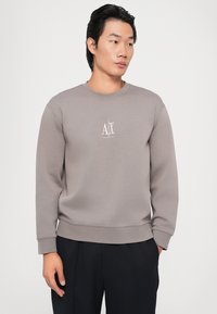 Felpa grigia con scollatura a girocollo, polsini e orlo a coste. Presenta un logo bianco sul petto. Il modello indossa pantaloni blu navy.