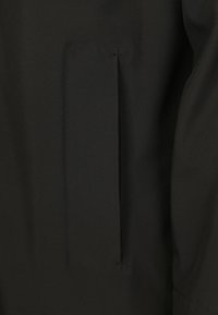 Zwart stof met een verticale splitzak, voorzien van twee kleine steken die de boven- en onderkant van de zakopening beveiligen.