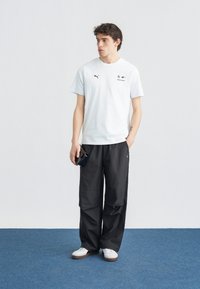 Νεαρός άνδρας που φοράει λευκό μπλουζάκι Puma Motorsport, μαύρο παντελόνι φαρδύ και λευκά αθλητικά παπούτσια, στέκεται σε μπλε χαλί, κρατώντας ένα μαύρο πορτοφόλι.