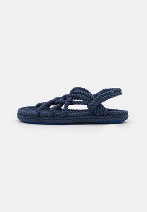 Marineblå sandal med sål og stropper lavet af snoet reb, med et løkkesløjfedesign og flad sål mod en hvid baggrund.