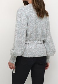 Femme portant un pull tricoté gris clair avec une ceinture nouée à la taille et un pantalon noir, vue de dos sur un fond uni.