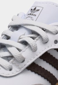 Λευκό αθλητικό παπούτσι Adidas με λευκά κορδόνια, μαύρες τρεις ρίγες και το λογότυπο Trefoil της Adidas στη γλώσσα, ορατό κοντά στην κορυφή.