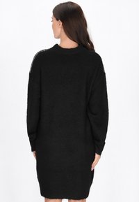 Zwarte gebreide sweaterjurk met een relaxte pasvorm, lange mouwen en een geribde halslijn, gemaakt van een zachte, textuurstof met een iets doorschijnend detail.