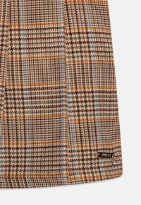 Brązowa i kremowa tkanina w kratkę z pomarańczowymi i niebieskimi akcentami, z delikatną teksturą i małą, metalową metką z logo po boku.