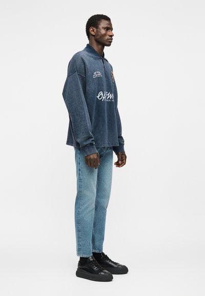 Άνδρας που στέκεται με μπλε γραφικό μακρυμάνικο πουκάμισο, ανοιχτόχρωμο μπλε τζιν και μαύρα αθλητικά παπούτσια μπροστά σε απλό λευκό φόντο.