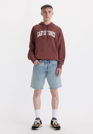 Brauner Kapuzenpullover mit dem Text "EARTH TONES", hellblaue Jeansshorts, weiße Socken und Sneakers in gemischten Farben. Schlichter Hintergrund.