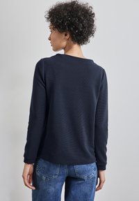 Mörkblå långärmad topp med struktur, lös passform och rund halsringning, tillsammans med ljusblå denimjeans och synlig ryggdetalj.