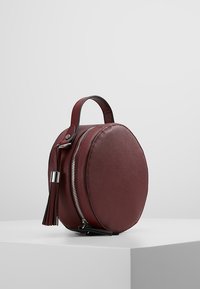 Rund bordeaux taske lavet af tekstureret materiale, med lynlåslukning, en sideskæft og et kort håndtag.