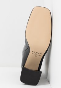 Semelle de chaussure en cuir noir avec talon carré, taille 37, marque "Jonak Paris", fabriquée au Portugal, positionnée à la verticale contre des surfaces blanches.