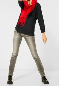 Svart oversized tröja, röd fransad scarf, glänsande silverfärgade slim-fit jeans och svarta snörstövlar mot en vit bakgrund.