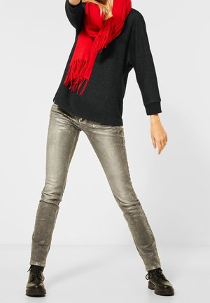 Czarny oversize sweter, czerwony szalik z frędzlami, błyszczące srebrne skinny jeans i czarne buty z wiązaniem na tle białym.