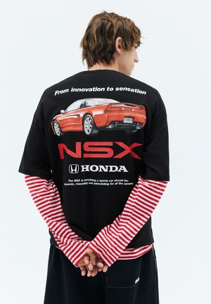 Person set bagfra bærer en sort Honda NSX T-shirt over en rød og hvid stribet langærmet shirt, hænderne foldet bag ryggen.