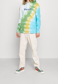 Sudadera con un estampado de tie-dye que presenta tonos azul, verde y amarillo, un bolsillo frontal y el logo "huf". Combinada con pantalones color crema y zapatillas.