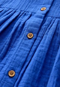 Tissu texturé bleu avec trois boutons marron à quatre trous en rangée verticale et des plis froncés le long de la couture.