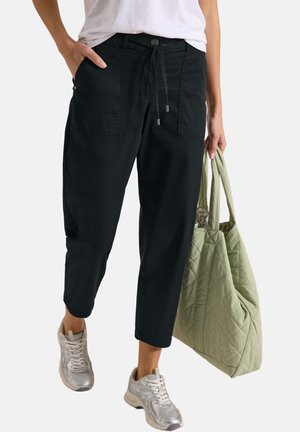 Frau trägt schwarze kurze Hose, weiße Sneakers und weißes Hemd und hält eine hellgrüne gesteppt tote Tasche in der linken Hand.