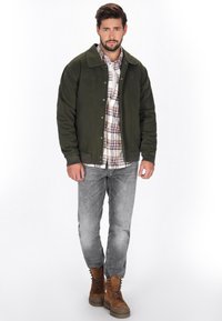 Mørkegrøn bomberjakke med knaplukning, broderede detaljer, båret over en ternet skjorte med knapper, parret med grå jeans og brune støvler.