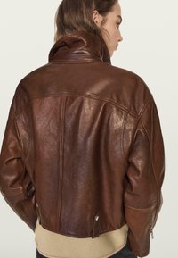 Braune Lederjacke mit kurzem Schnitt, steigendem Kragen und seitlichen Reißverschlüssen. Glatte Textur mit sichtbaren Nähten und dezenten Taschen.