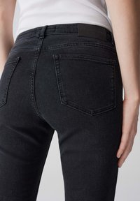 Jean en denim noir au design ajusté, avec deux poches arrière et une texture lisse, vu de côté. Étiquette de taille mate visible.