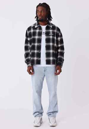 Homme aux cheveux tressés portant une chemise à carreaux noir et blanc par-dessus un t-shirt blanc imprimé, un jean bleu clair et des baskets blanches, debout.