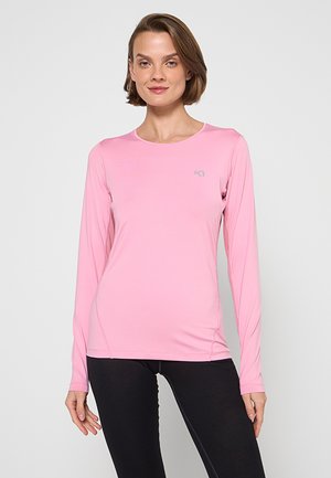 Donna in piedi che indossa una maglia atletica aderente a maniche lunghe rosa e leggings neri, rivolta in avanti con un'espressione neutra.