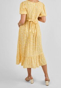 Frau in einem gelben Blumen-Midikleid mit Puffärmeln und gebundenem Taillengürtel, von hinten gezeigt, kombiniert mit goldenen Blockabsatzsandalen.