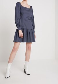 Vestido de lunares marino con mangas abullonadas, escote cuadrado y dobladillo con volantes. Combinado con botas de tacón blancas para un look moderno.