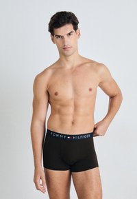 Braune Boxershorts mit einem marineblauen Bund, der das Logo „TOMMY HILFIGER“ trägt. Glatte Textur und enganliegendes Design. Modell steht mit einer Hand auf der Hüfte.