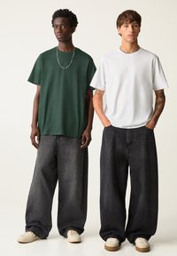Two individuals wearing oversized t-shirts; one in dark green, the other in white. Both have baggy dark jeans and casual shoes.