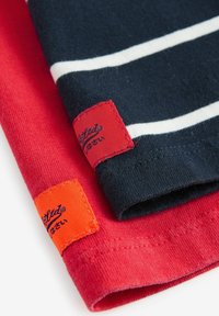 Zwei Baumwoll-T-Shirts, eines rot und eines marineblau mit weißen Streifen, mit kontrastierenden Markentags in Rot und Orange und sichtbaren Nähdetails.