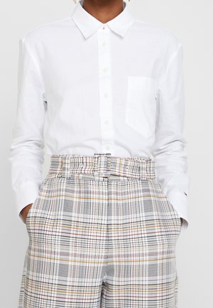 Persoon draagt een wit overhemd met knopen, ingestopt in een hooggetailleerde, geruite broek met riem, met handen in de zakken.