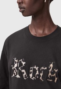 Musta puuvillapaita, jossa on suuri, tekstuuri "Rock" -grafiikka harmaa-beige leopardikuosisella fontilla. Nesti kaula-aukko.