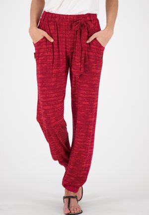 Woman wearing loose red patterned pants with an elastic waist, side pockets, tied belt, and black sandals against a white background.