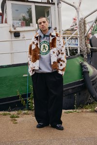 Mladý muž oblečený ve skvrnitém hnědo-bílém kožichu přes mikinu Oakland Athletics stojí na nábřeží poblíž zelené lodi.
