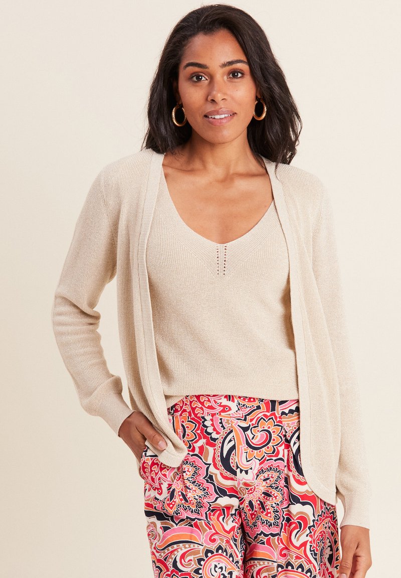 Cardigan beige chiaro indossato sopra una maglietta a scollo a V di colore chiaro con piccoli dettagli, abbinato a pantaloni dai vivaci motivi floreali nelle tonalità di rosa e rosso.