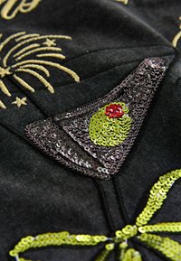 Czarna tkanina z misternym wzorem z cekinów przedstawiającym kieliszek do koktajlu z zieloną oliwką i czerwonym akcentem, ozdobiona złotym niciowym detalem.
