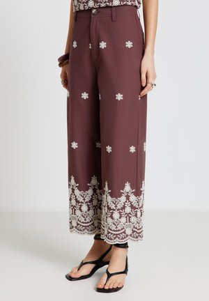 Pantaloni marroni a gamba larga con ricamo floreale bianco sull'orlo e sparso in tutto, indossati con sandali neri con cinturino alla caviglia.