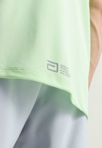 Camiseta atlética verde claro con un tejido texturizado. Presenta un logo impreso en gris cerca del dobladillo. Combinada con pantalones cortos gris claro.