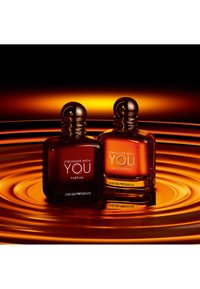Twee parfumflessen op een golvende oranje achtergrond. De linkerfles is donker amberkleurig met een ronde dop; de rechter is lichter, ook rond, met witte tekst.