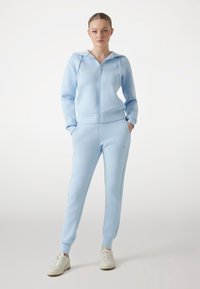 Light blue zip-up hoodie and matching joggers in soft fabric; features ribbed cuffs and a hood. Paired with white sneakers.