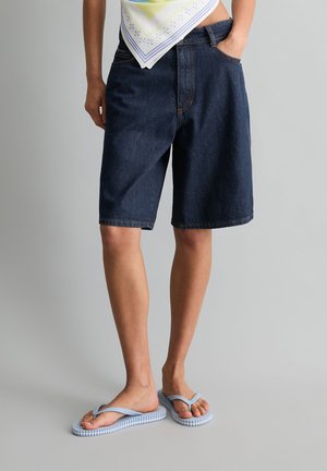 Persoon draagt donkerblauwe denim Bermuda shorts, een lichtgekleurde top met bandana-patroon en lichtblauwe teenslippers, staand met één hand in de zak.