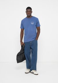 Blue cotton t-shirt with "OBEY" logo on the chest, paired with loose-fitting blue jeans and white sneakers, holding a black jacket.