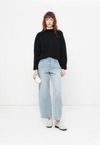 Crni oversized pulover, svijetloplave hlače širokih nogavica i bijele slip-on cipele. Model drži malu bijelu torbicu. Minimalistički stil, neutralne boje.