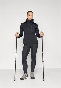 Black softshell jacket with a zippered chest pocket, paired with fitted dark leggings and hiking shoes, held hiking poles in each hand.