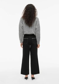 Sivi teksturirani pulover, široke crne traperice s stražnjim džepovima prošivenim malim metalnim studovima, i crne cipele na visoku petu sa špicastim vrhom. Vidljive kovrče na stražnjoj strani.