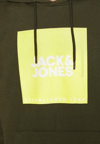 Sudadera color verde oliva con un gran gráfico cuadrado amarillo que presenta el texto blanco "JACK & JONES" y "ESTABLECIDO EN 1990" en la parte inferior.