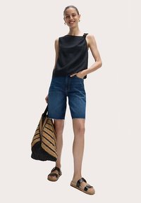 Top nero senza maniche con nodo laterale, pantaloncini di denim blu, borsa tote intrecciata nera e beige, e sandali neri con suola spessa.