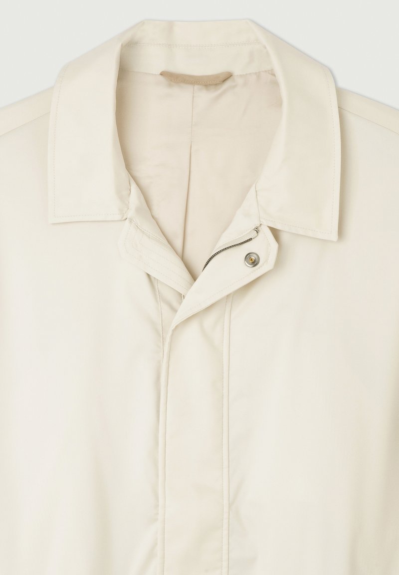 Manteau beige clair avec un col classique, une fermeture éclair avec un bouton pression et une texture de tissu lisse. Design minimaliste avec des coutures subtiles.