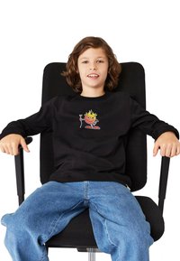 Mustat pitkät hihat -paita, jossa on värikäs grafiikka hahmosta, yhdistettynä vaaleansiniset farkkushortsit, istuu mustassa toimistotuolissa.