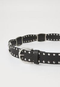 Schwarzer perforierter Ledergürtel mit einer silberfarbenen Schnalle, größeren Löchern und Akzenten entlang der Länge, die einen strukturierten Look bieten.