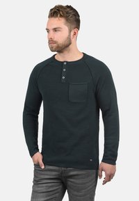 Donkergroene longsleeve shirt met een gestructureerde oppervlakte, ronde hals, drie knopen en een borstzak. Gemaakt in combinatie met grijze jeans.